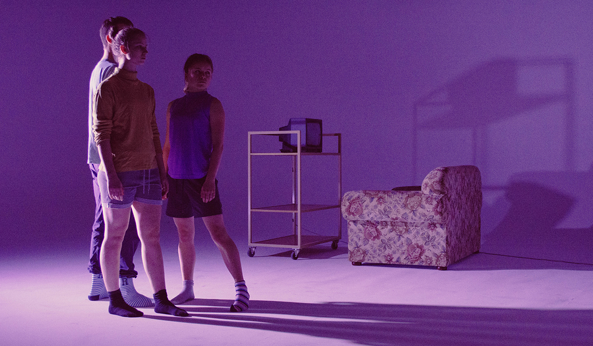 Three people standing in a purple lit room with 1 couch and a tv.